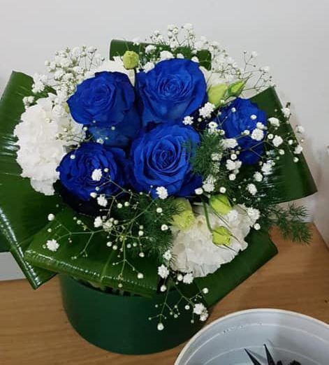Composizione floreale: rose blu, garofani bianchi, gypsophila, foglie verdi, in un contenitore verde.