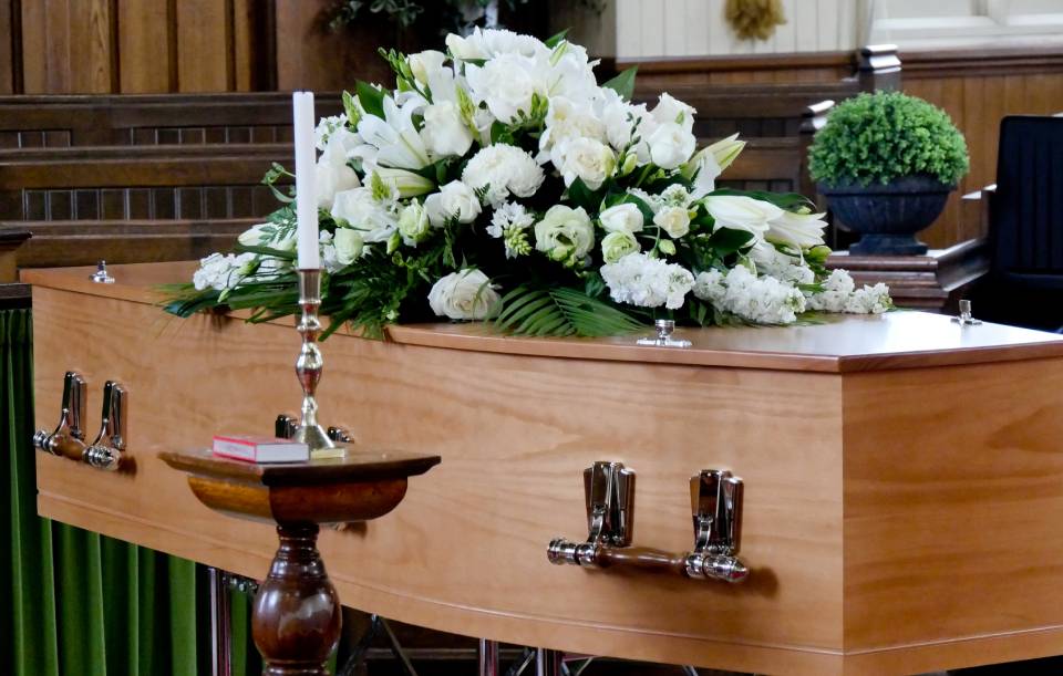 Una bara di legno con una composizione floreale bianca, una candela e un tavolino in un ambiente da chiesa.