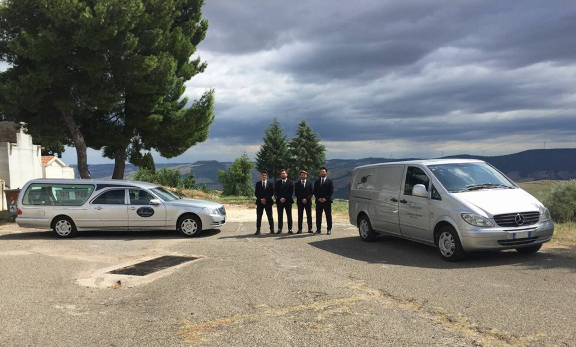Cinque persone in abiti scuri in piedi tra un carro funebre e un furgone su una strada che domina un paesaggio.