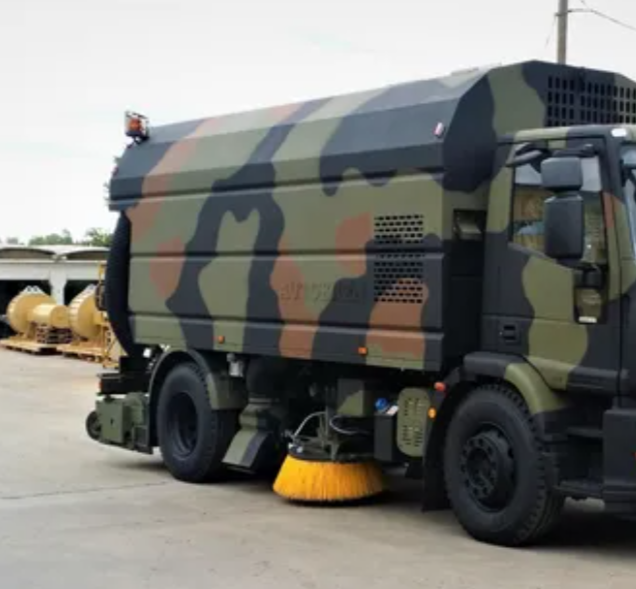 Camion spazzaneve con verniciatura mimetica, dotato di una grande spazzola, parcheggiato sul marciapiede.