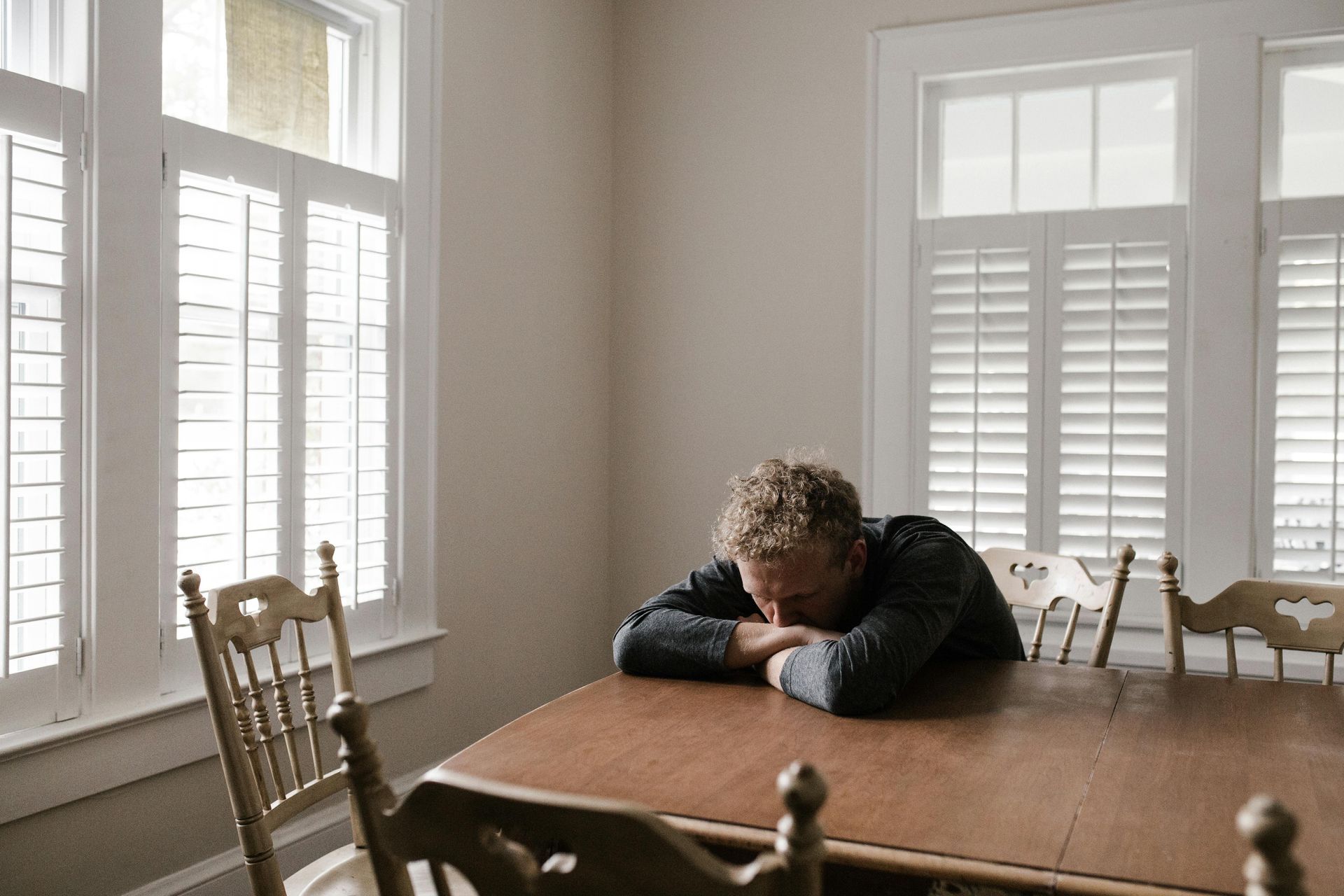 Man with head on table in a room with shuttered windows, looking dejected.