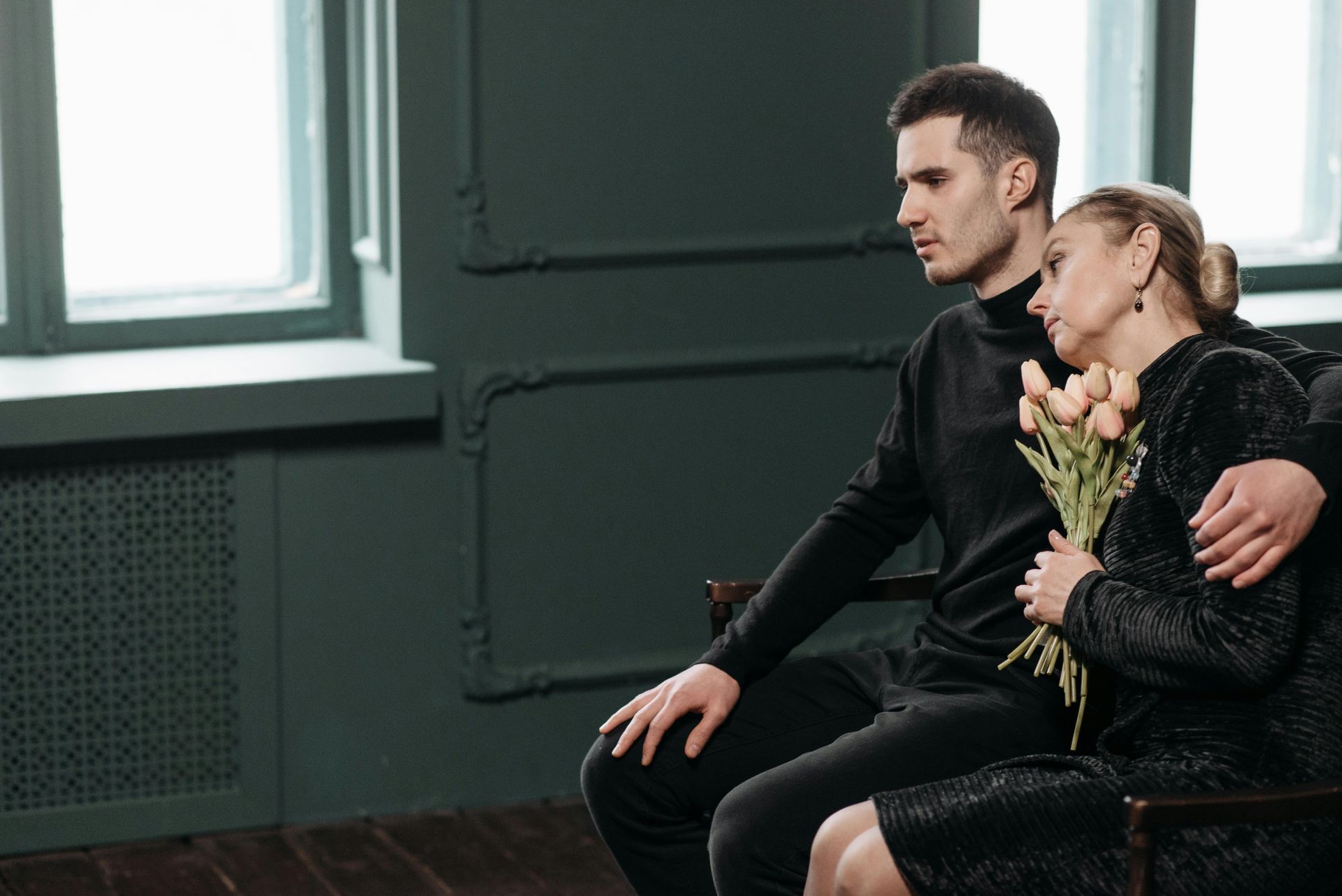 Man comforts woman, both in black, holding flowers, seated near a window in a dark room.