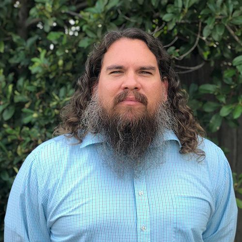 A man with long hair and a beard is wearing a blue shirt and standing in front of a tree.