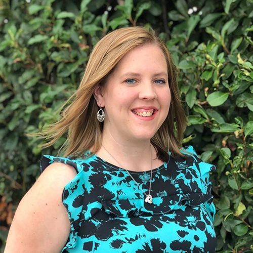 A woman in a blue and black dress is smiling in front of a bush.