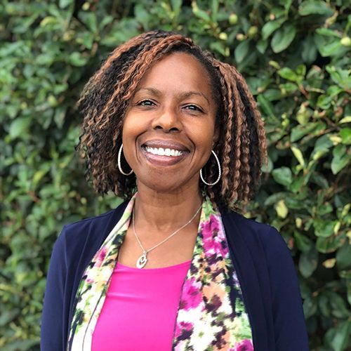 A woman with dreadlocks is smiling for the camera in front of a bush.