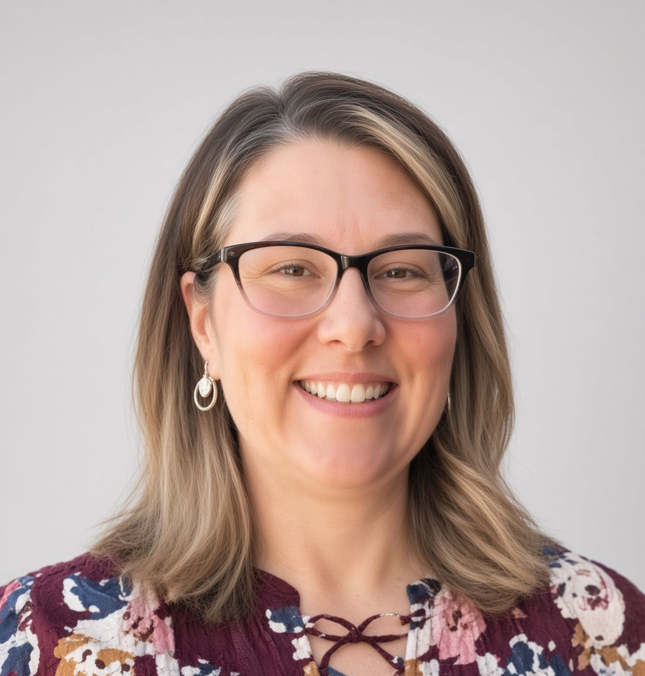 A woman wearing glasses and a floral shirt is smiling in front of a brick wall.