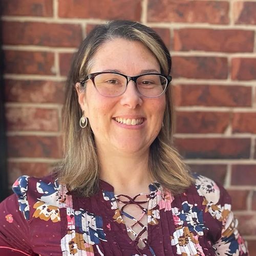 A woman wearing glasses and a floral shirt is smiling in front of a brick wall.