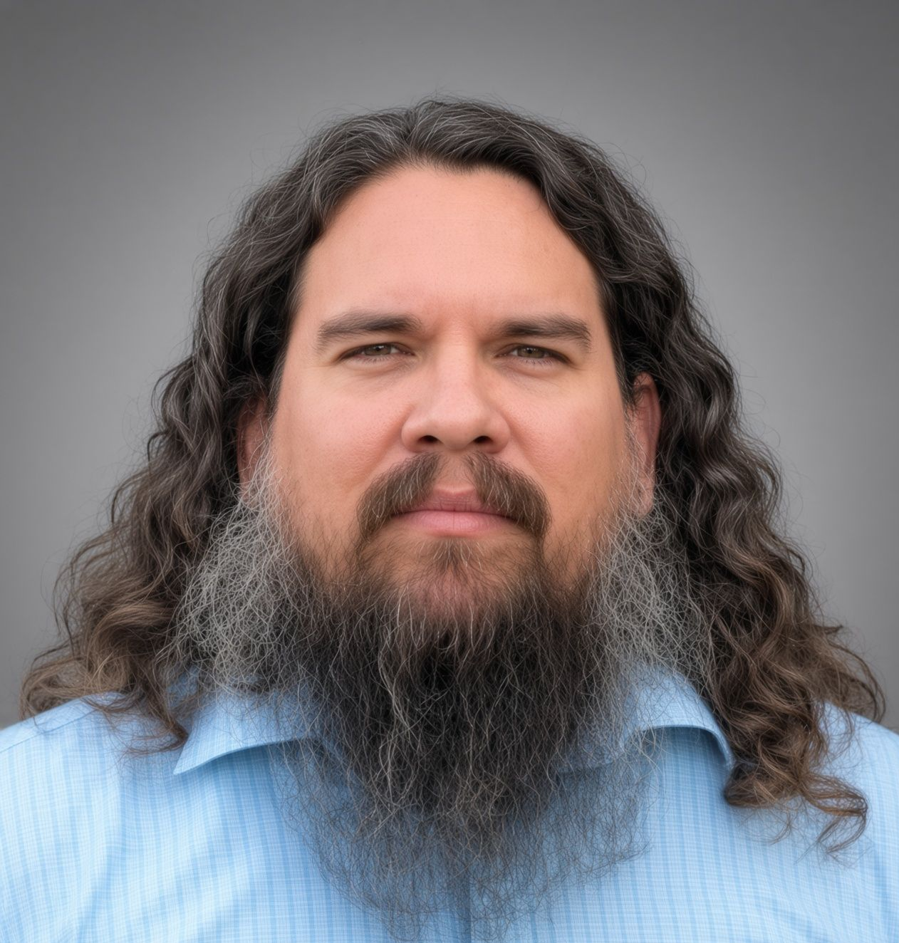A man with long hair and a beard is wearing a blue shirt and standing in front of a tree.