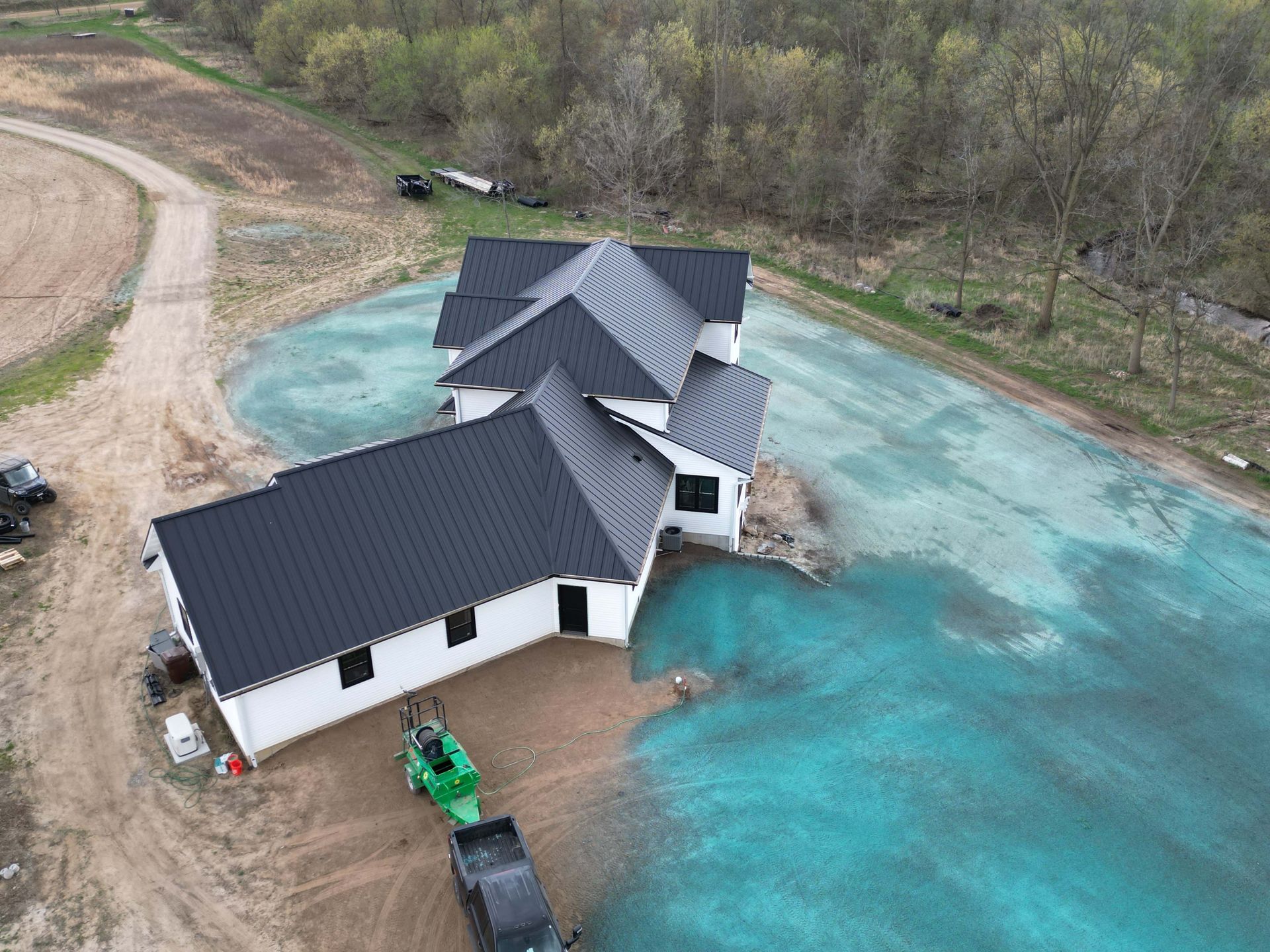 Aerial view of a white house with a black roof, near a dirt road and blue-green grass, a tractor.