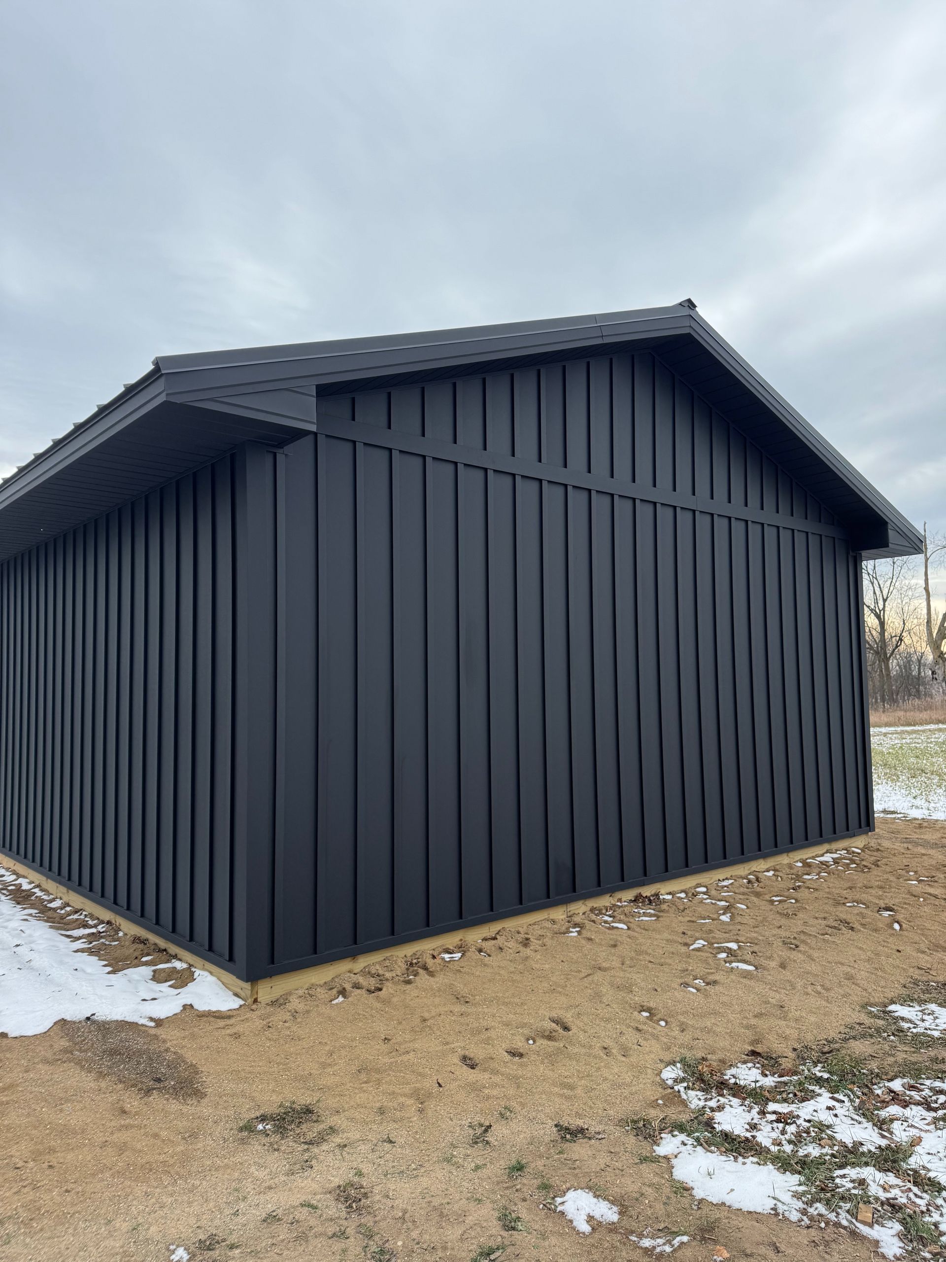 Black-sided shed with dark shingle roof, on a gravel bed with patches of snow, against a cloudy sky.