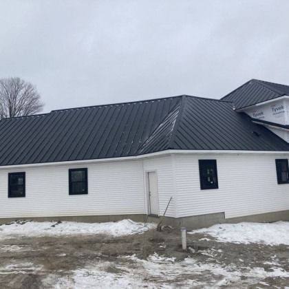 White building with black metal roof and windows; construction site, snowy ground.