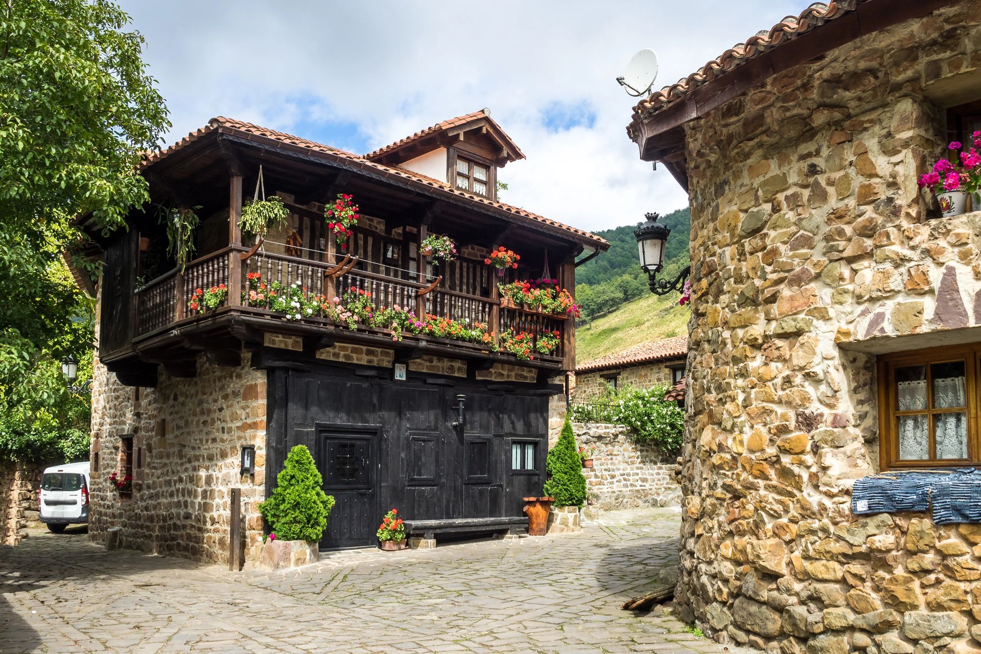 Edifici in pietra in un villaggio con un balcone pieno di fiori e una strada acciottolata.