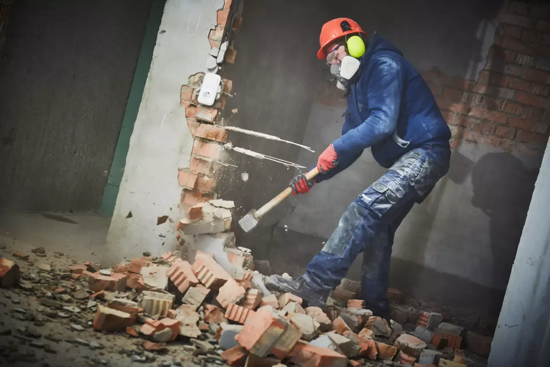 Un operaio edile che demolisce un muro di mattoni, indossando dispositivi di sicurezza, all'interno di un edificio.