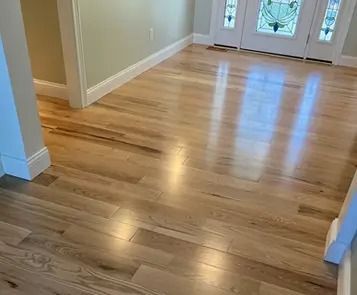 Light-colored hardwood floor in a well-lit entryway, reflecting light. White trim and a partially visible door.