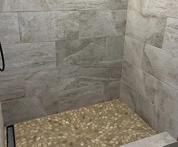 Shower stall with gray tile walls and a pebble-stone floor; black drain on the bottom.