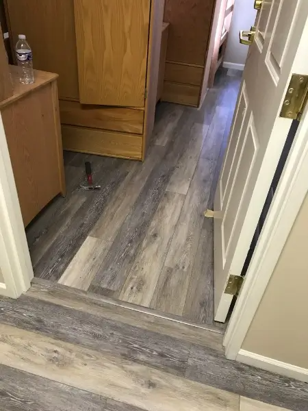 Gray wood-look flooring installed in a hallway with light-colored walls and wood furniture. A door is open.
