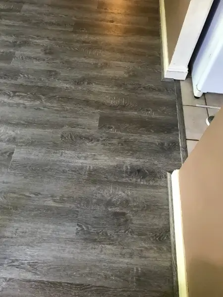 Dark gray wood-look flooring transitions to light tile in a kitchen. A cream-colored cabinet edge and wall are visible.