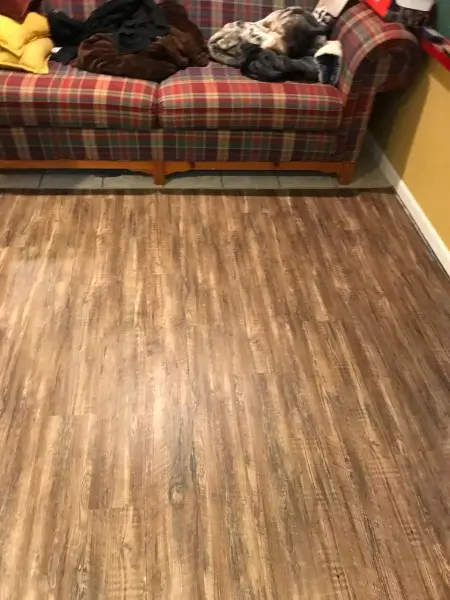 Brown wood-look flooring extends from the bottom of a plaid couch. Clothes are on the couch. Yellow wall is on the right.