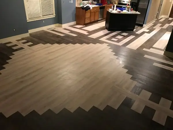 Wooden floor with geometric patterns in light and dark wood tones. Kitchen in the background.