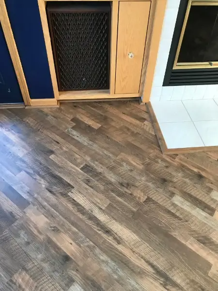 Wooden floorboards angled in a herringbone pattern next to a fireplace and a cabinet with a vent.