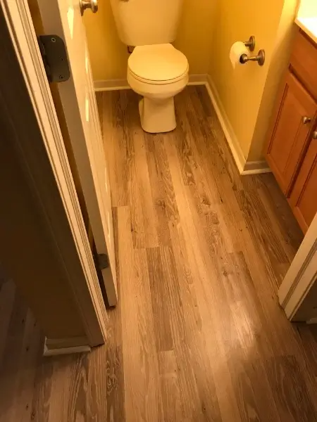 Bathroom with wood-look flooring. Toilet and wooden vanity are visible. Door partially open to the left. Yellow walls.