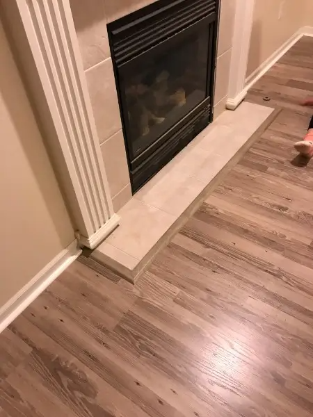 Fireplace with gray hearth, beige and black tile, set against wood-look flooring and beige wall.