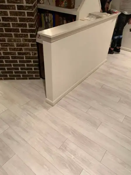White wooden-look flooring and a white wall with trim in an interior setting. A brick wall is visible to the left.