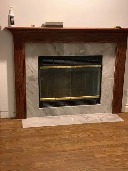 Fireplace with wooden mantel, marble-look tile surround, and glass front, set against a wood-paneled wall.