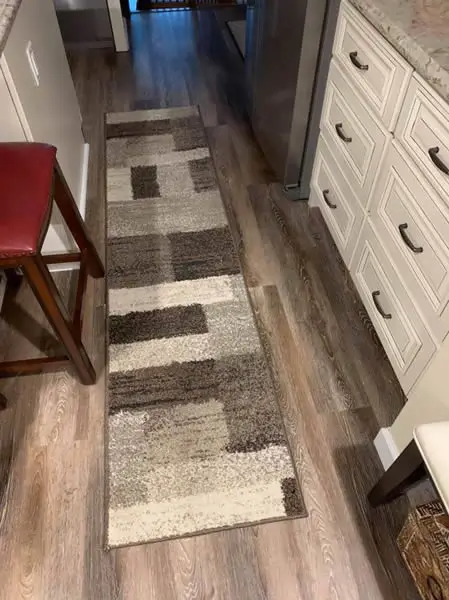 Narrow rectangular rug on a wood floor in a kitchen with white cabinets, a stool, and a refrigerator.