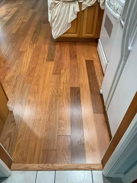 Wooden flooring in a kitchen, with various shades of brown. A white stove and cabinets are also visible.