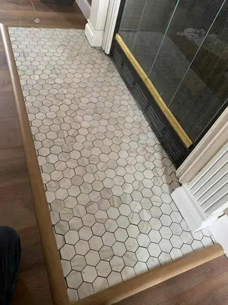 Fireplace hearth with hexagonal white tiles, bordered by brown wood trim, beside a dark fireplace.