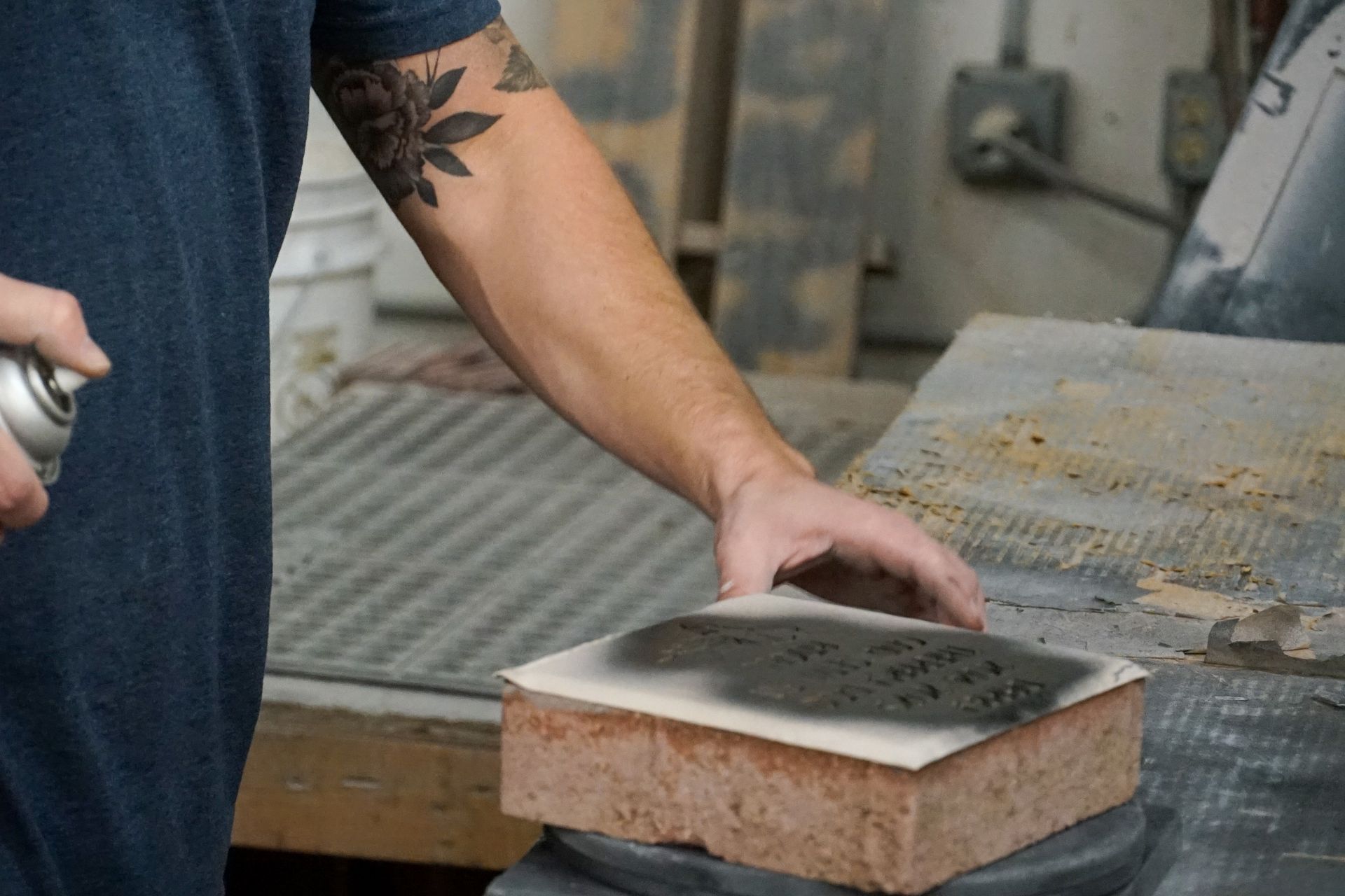 Person sprays dark paint onto a stencil on a brick in a workshop setting.