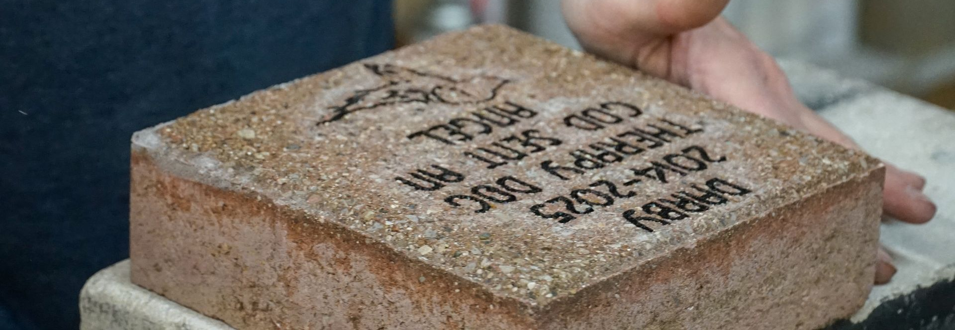 A person's hands holding a brick with engraved text. The brick is light brown with dark text.