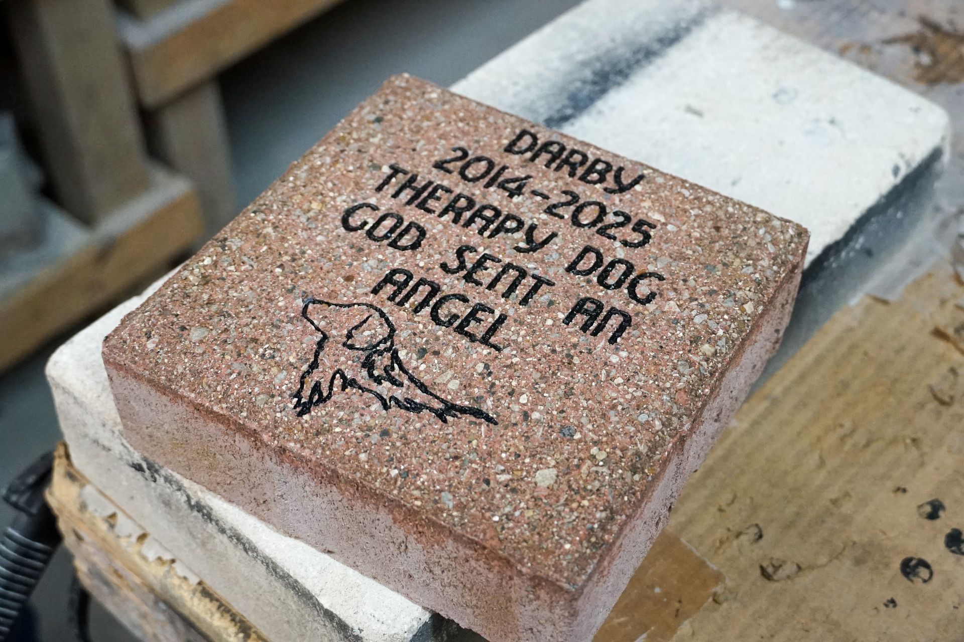 Memorial brick for a dog, with engraved name, dates, and an image of a dog; resting on other bricks.