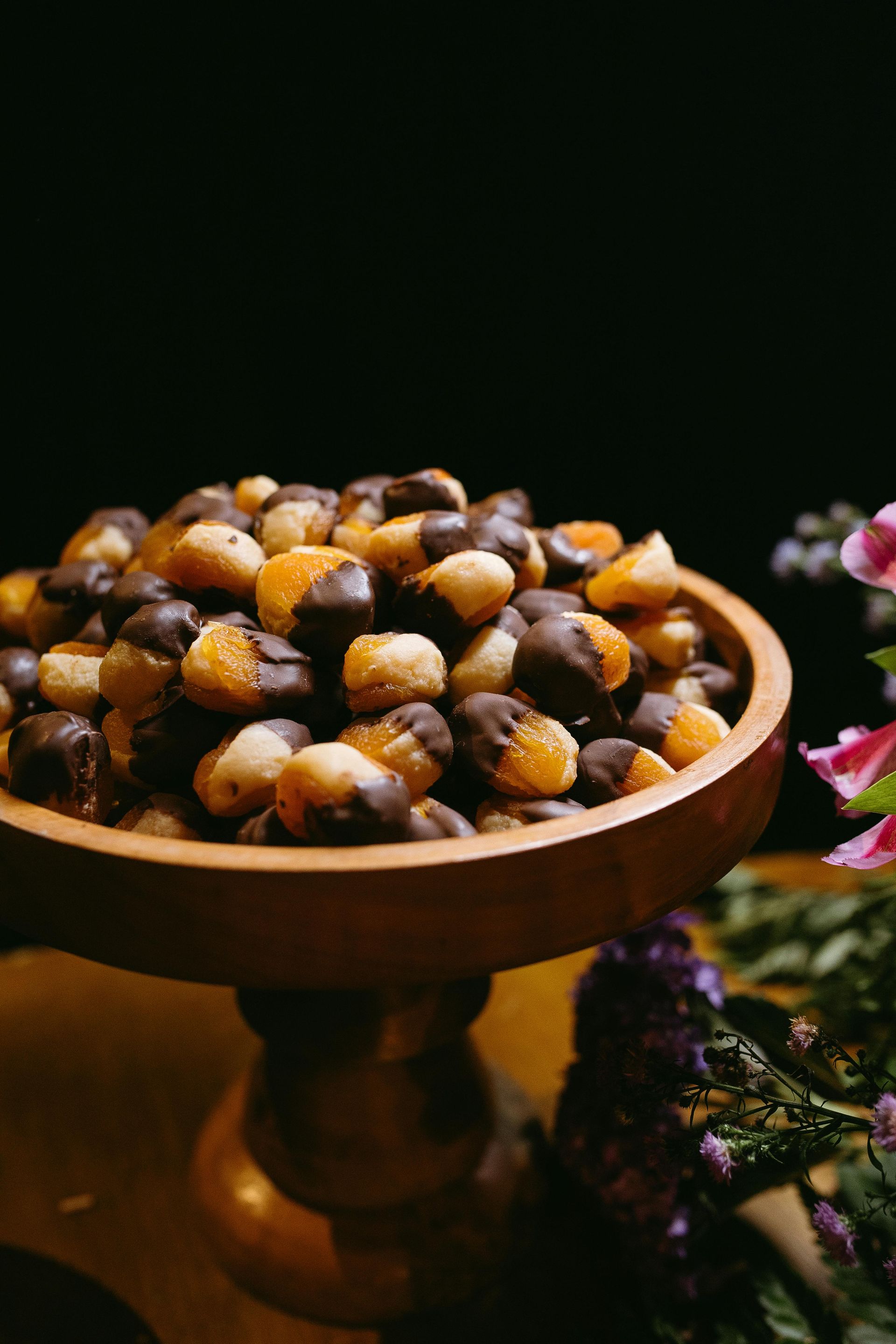 Wooden bowl of mixed sweets and nuts on a pedestal, with flowers in the background.