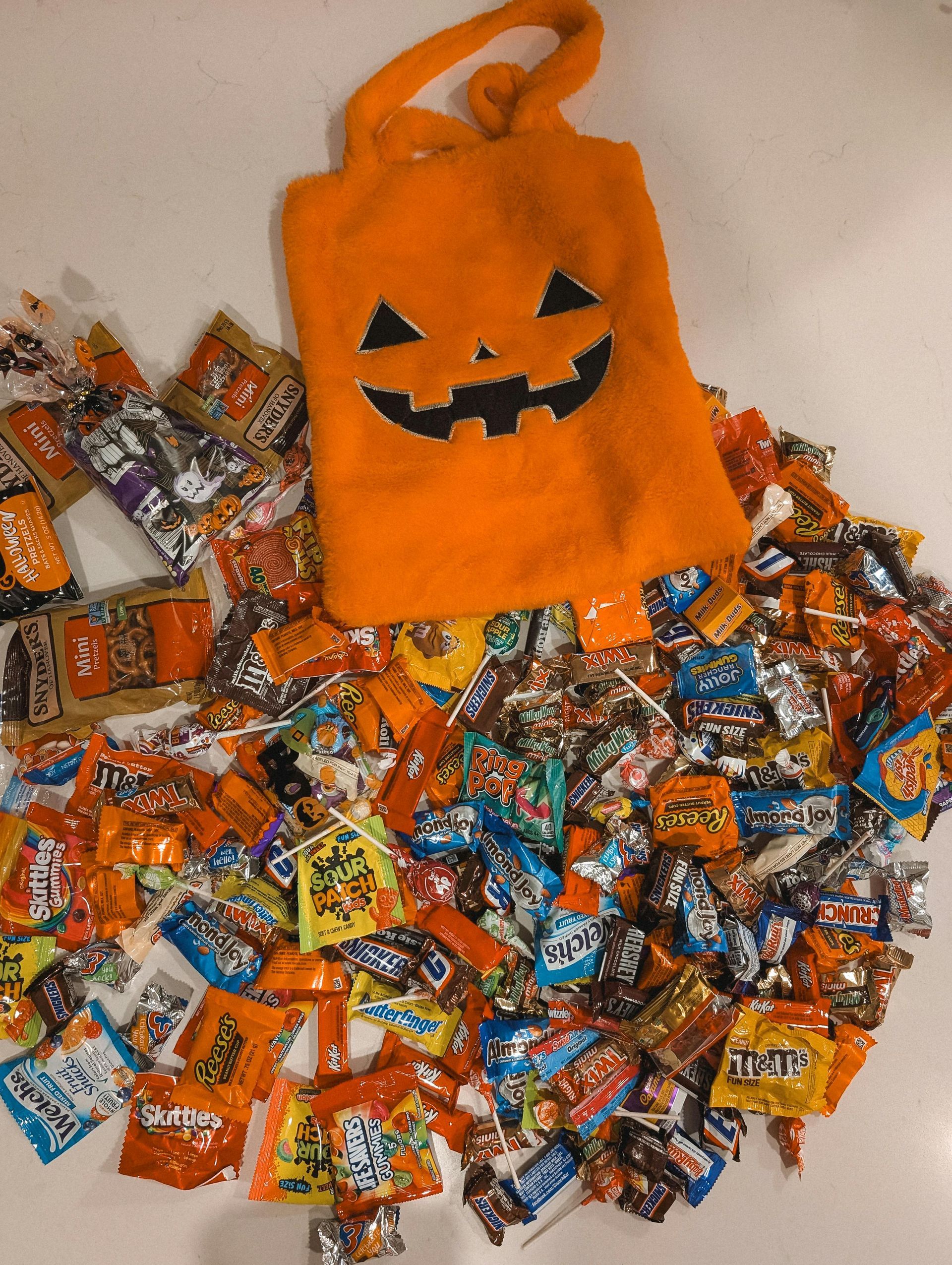 Orange Halloween candy bag with jack-o'-lantern face, surrounded by assorted wrapped candies