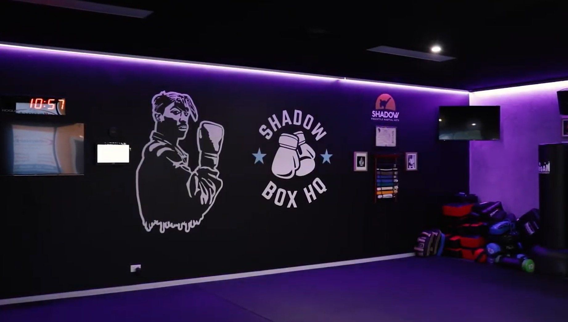 Boxing gym interior with purple lighting, logo on a black wall, and exercise equipment.