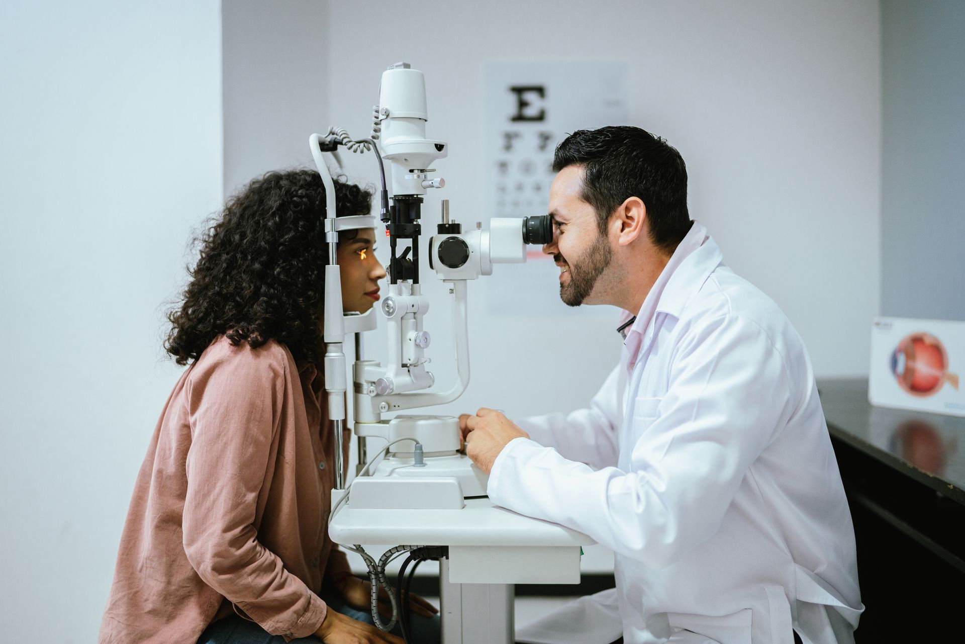 An optometrist, in a white uniform, is performing an optical exam on a young woman at a clinic.
