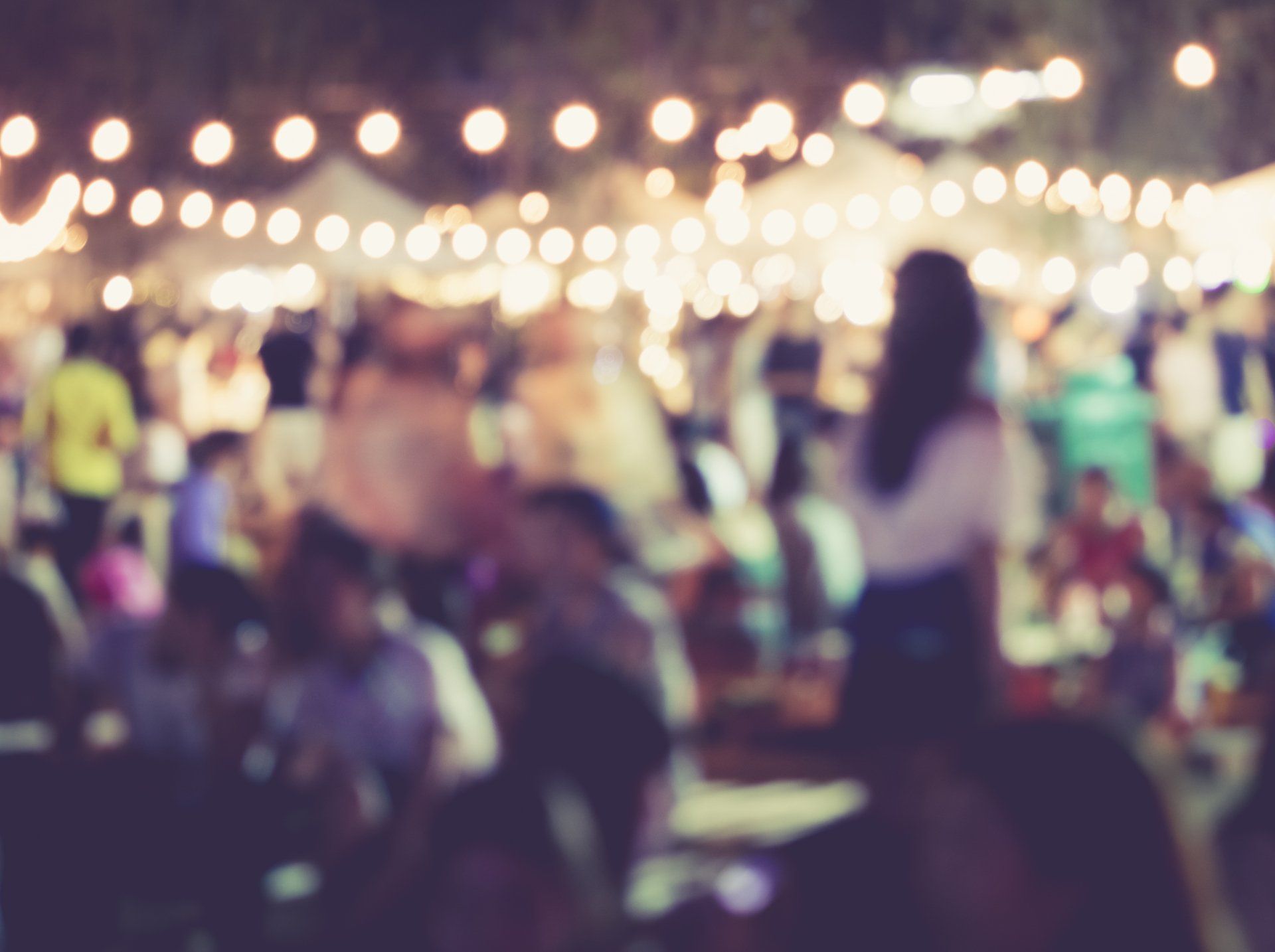 Blurred nighttime crowd at an outdoor market with string lights above. People are socializing near vendor tents.