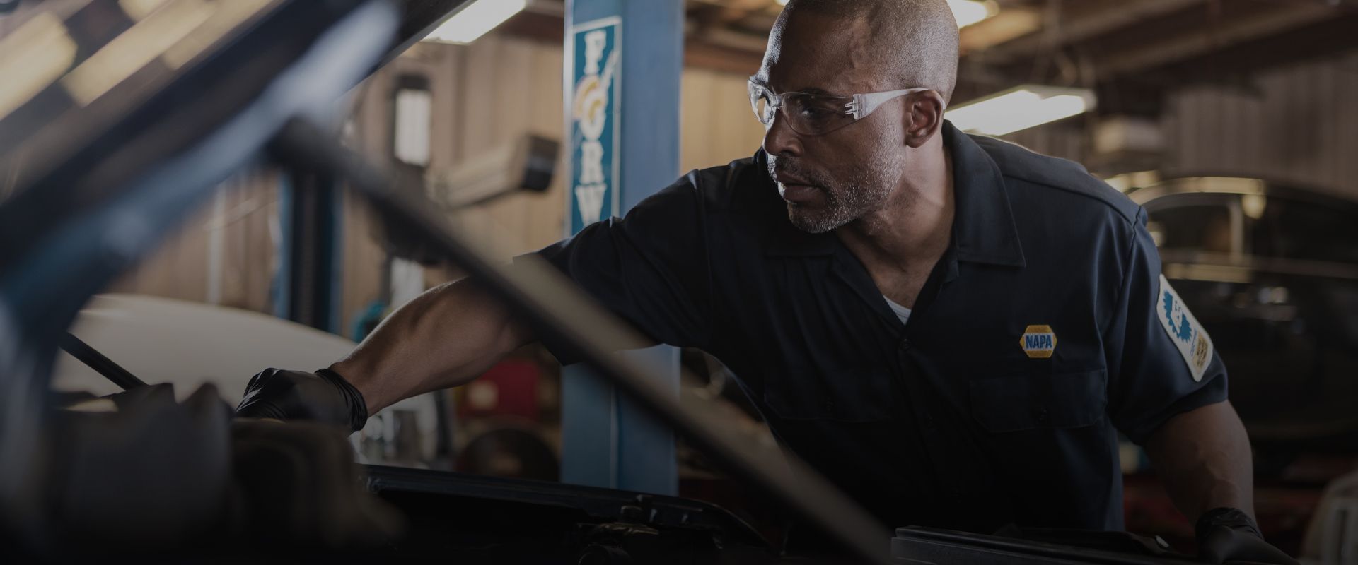 A mechanic in a navy shirt with the Napa logo looks at a car engine, wearing safety glasses. | Emge's Car Craft Auto Services