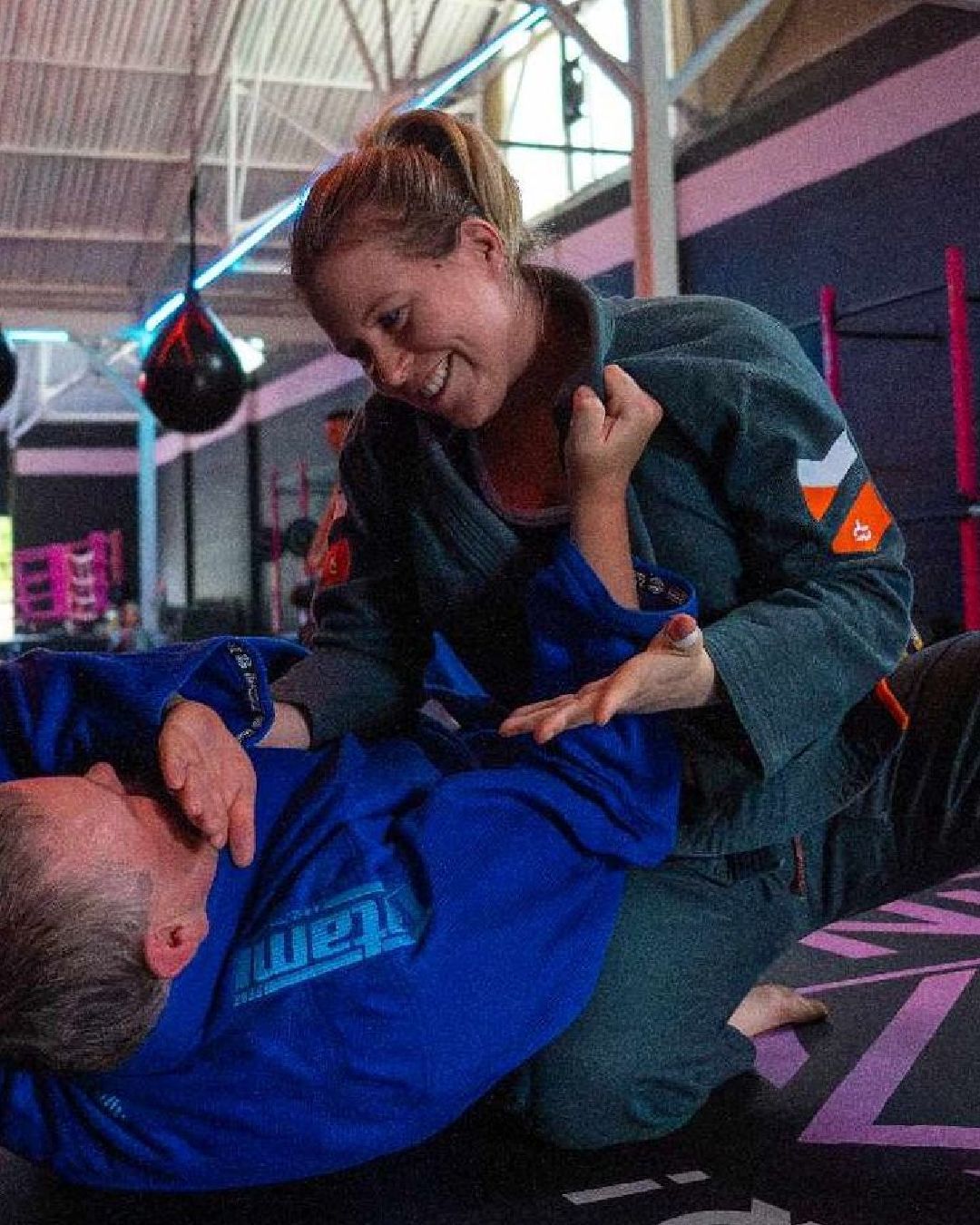 Two martial artists grapple on a mat in a gym, one in a blue gi and one in a dark gi.