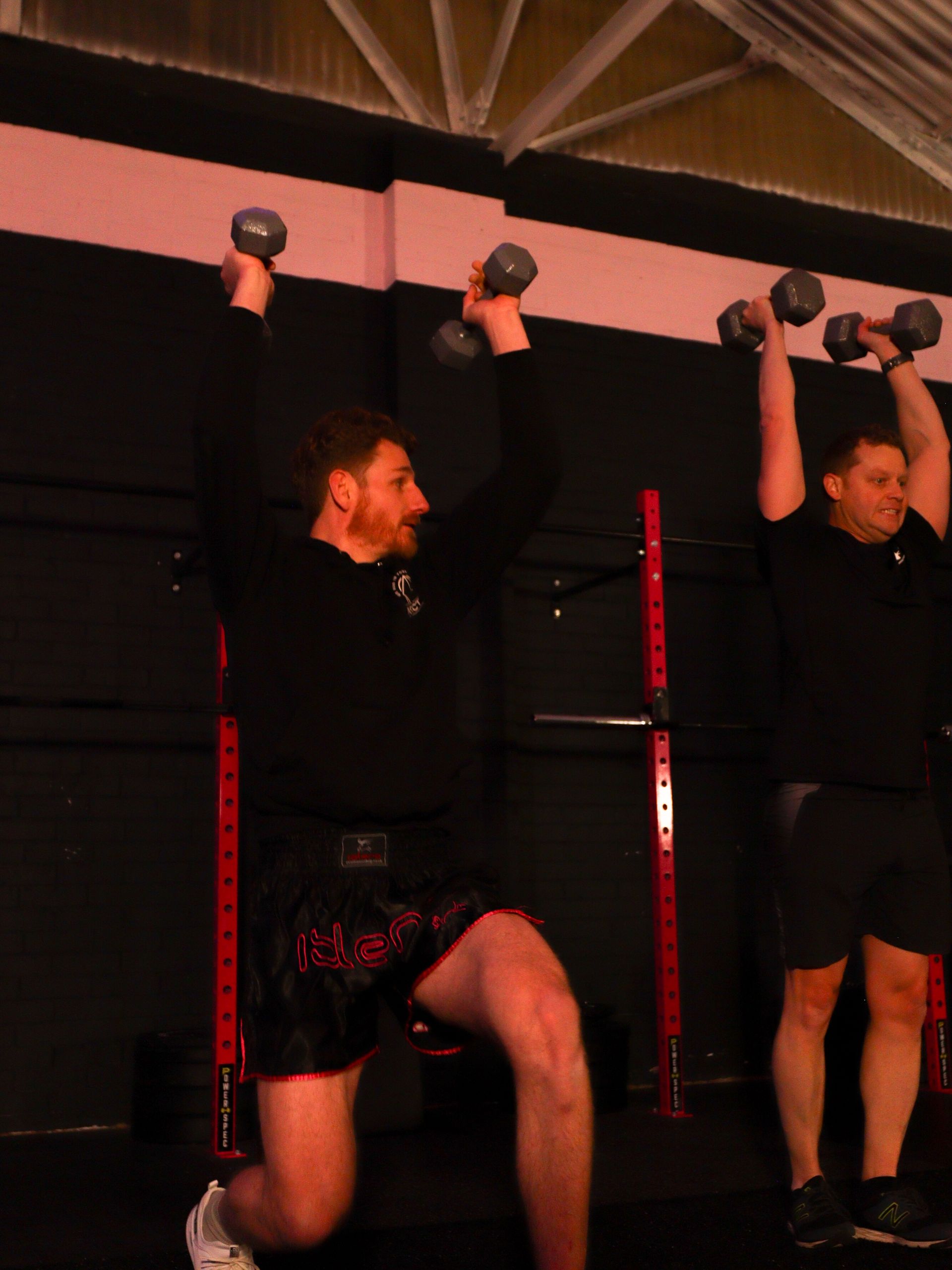 Two people doing wall squats indoors, holding dumbbells under a black ceiling with red rails.