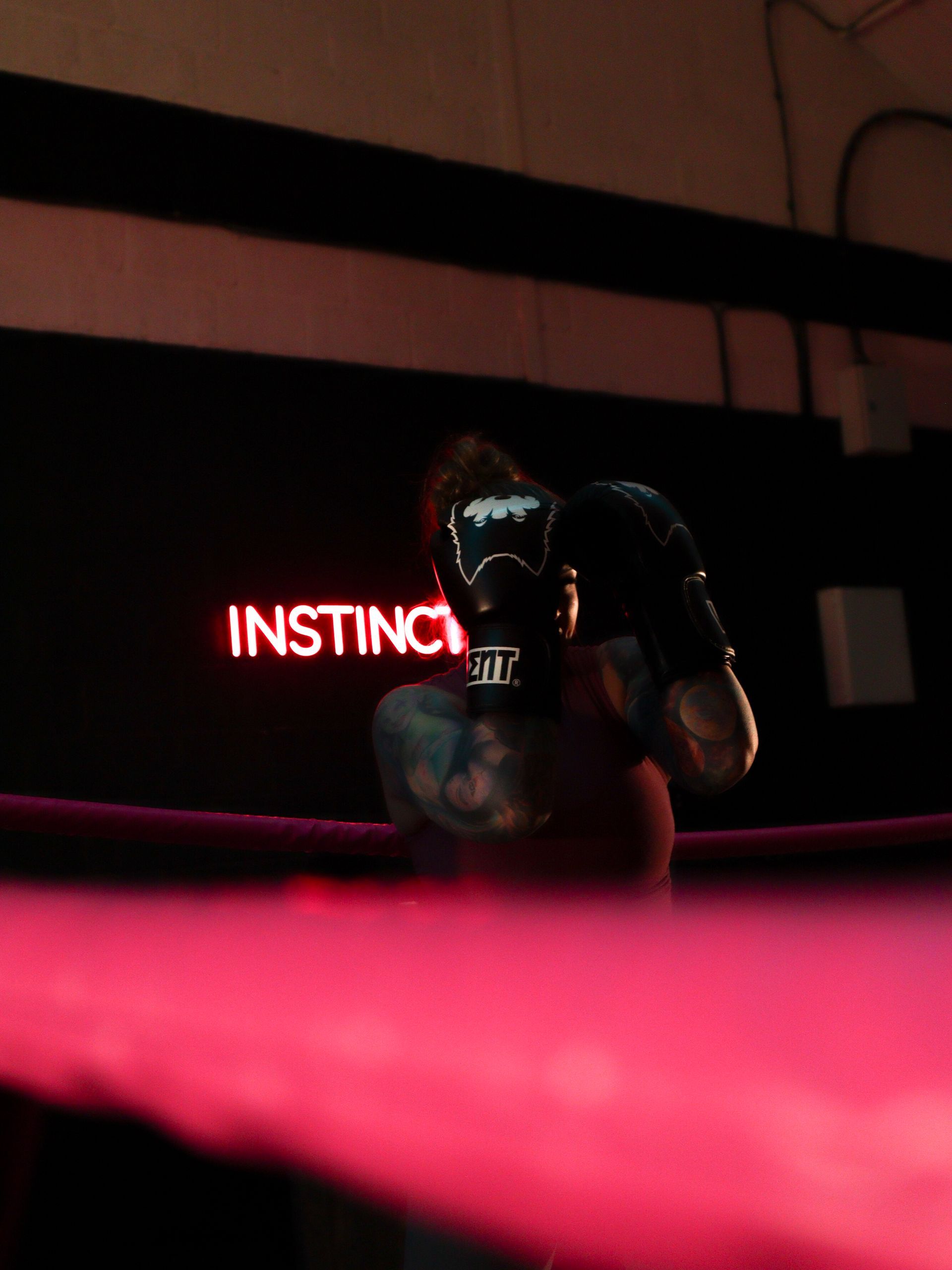 Gloved person crouches in a dark room, lit by red neon “INSTINCT” sign and pink foreground blur.