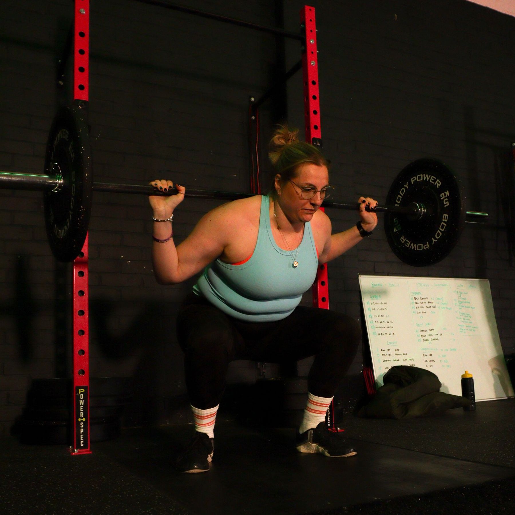 Woman performing a back squat with a barbell in a gym, wearing a turquoise top and black shorts.
