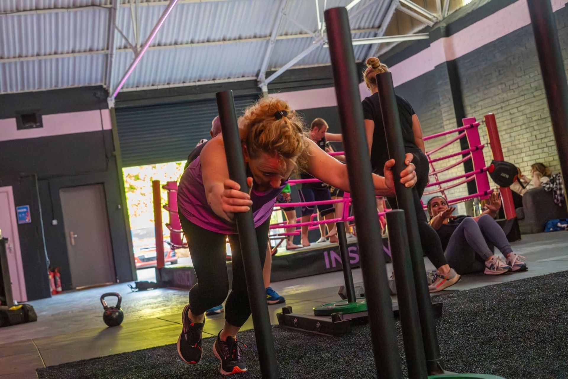 Person using a sled push machine in a gym, with pink railings and other people exercising nearby.
