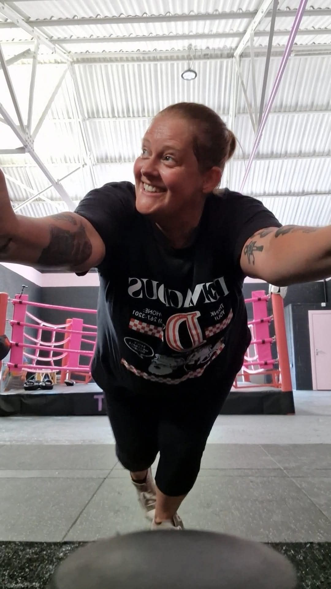 Smiling person leaning forward in a gym, holding arms out toward the camera under bright overhead lights