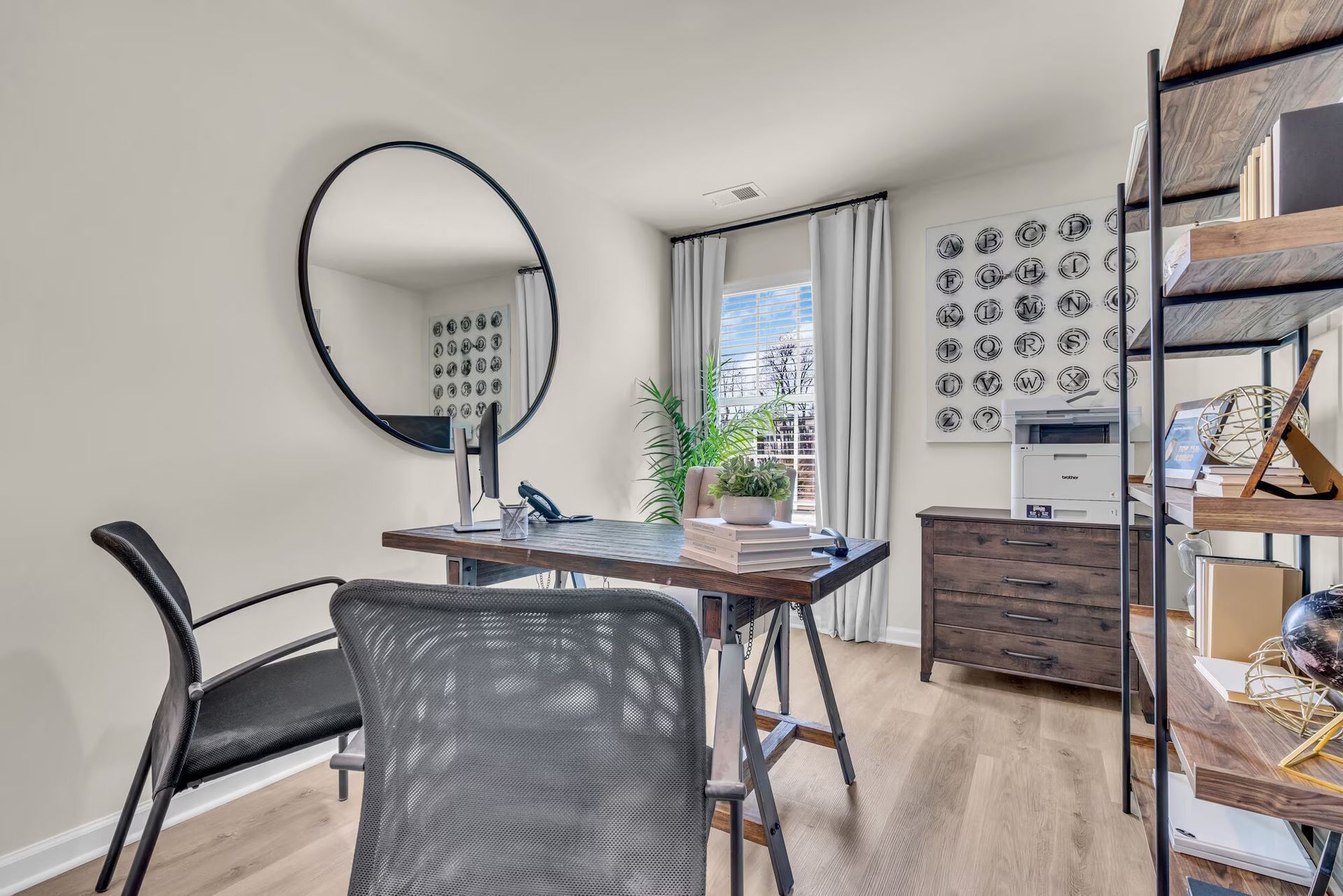 Modern home office with glass desk, black chair, round mirror, and shelving on a light wood floor.