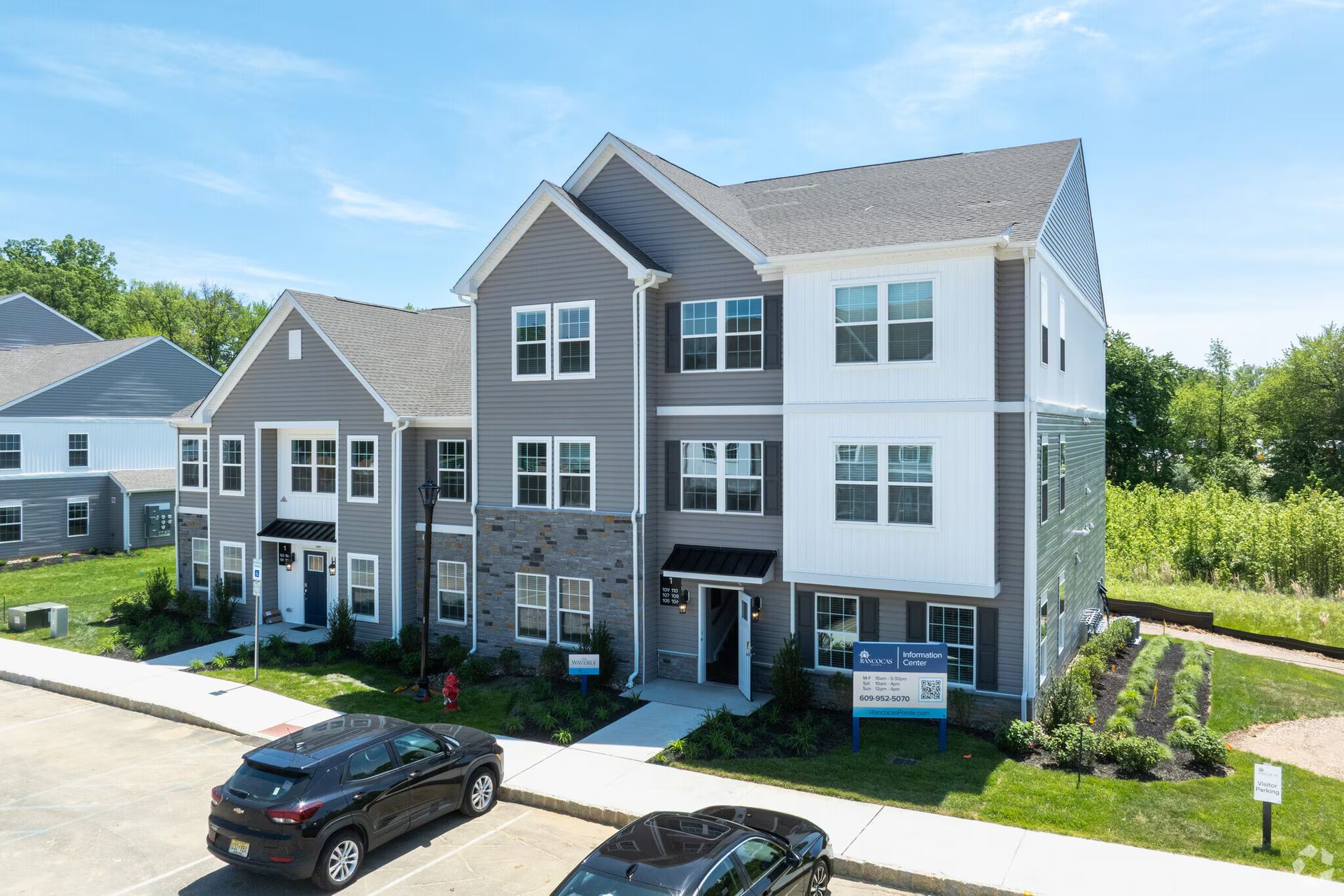 Modern apartment building with gray and white siding, parked cars, and a real estate sign out front