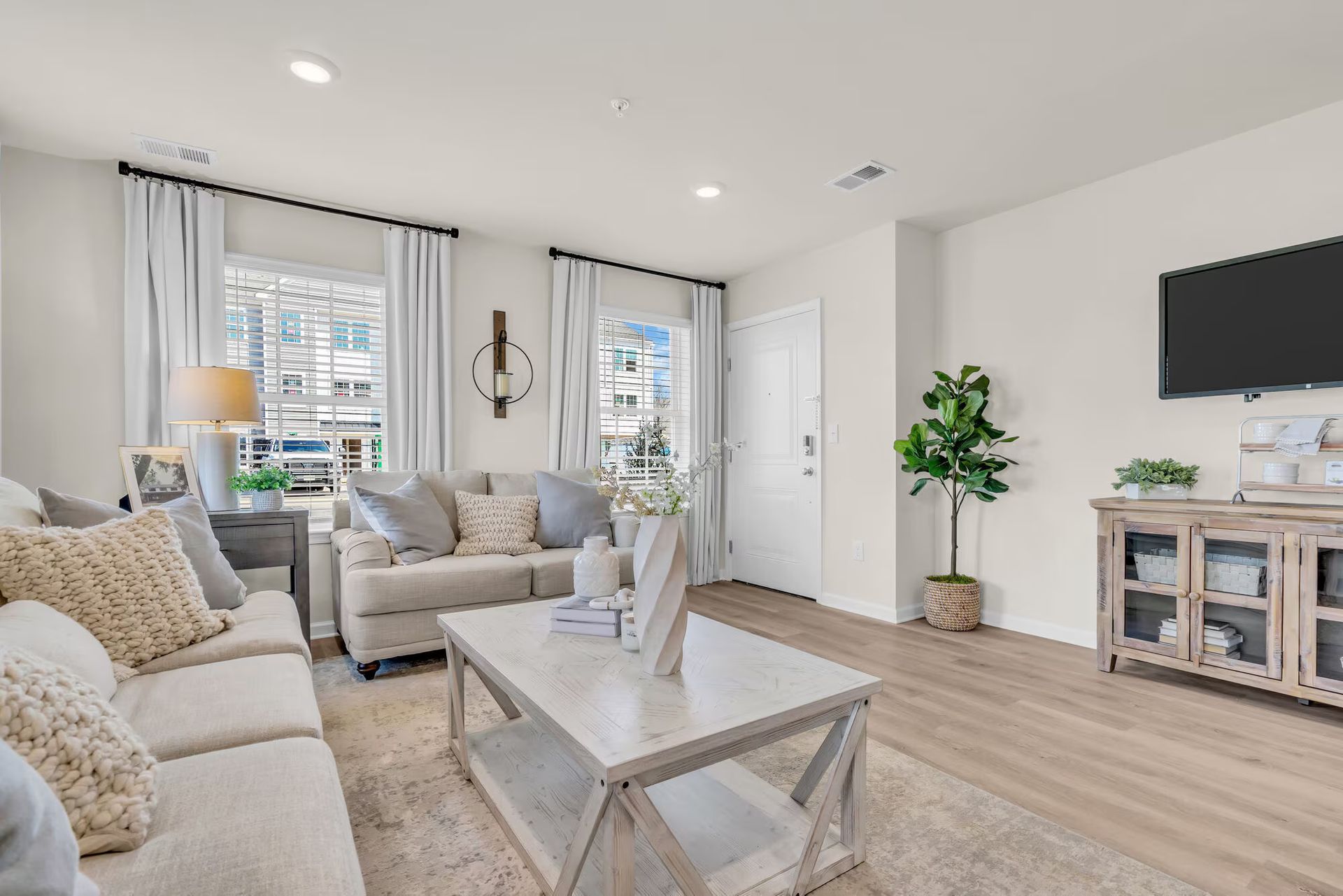 Bright living room with beige sofas, white coffee table, wall TV, and large windows