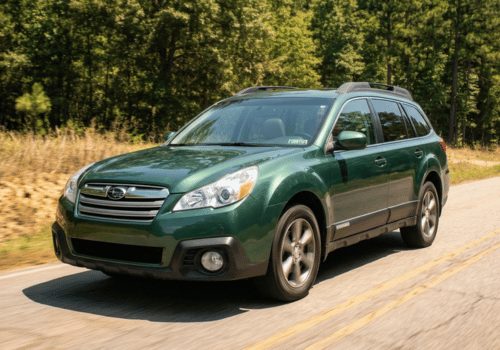 Green Subaru Outback driving along a two-lane road bordered by trees on a sunny day.